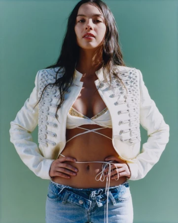 Portrait of a young adult female model in an off-white jacket with embellishments, light blue jeans, and a bikini top against a green backdrop.