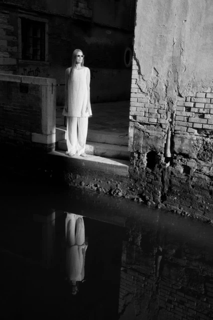 Monochrome shot of a female model in white clothing standing by a canal in Venice, Italy, reflecting in the water.