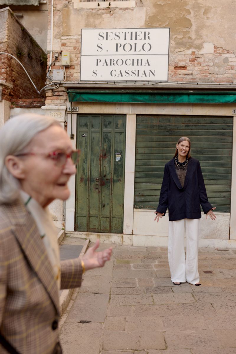 Two senior women pose on a brick street in Venice, Italy. One woman is smiling, and the other is out of focus in the foreground.