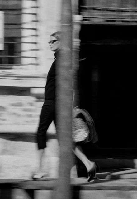Blurred monochrome shot of a stylish woman in sunglasses walking on a city street, holding a patterned handbag with an open doorway behind her.