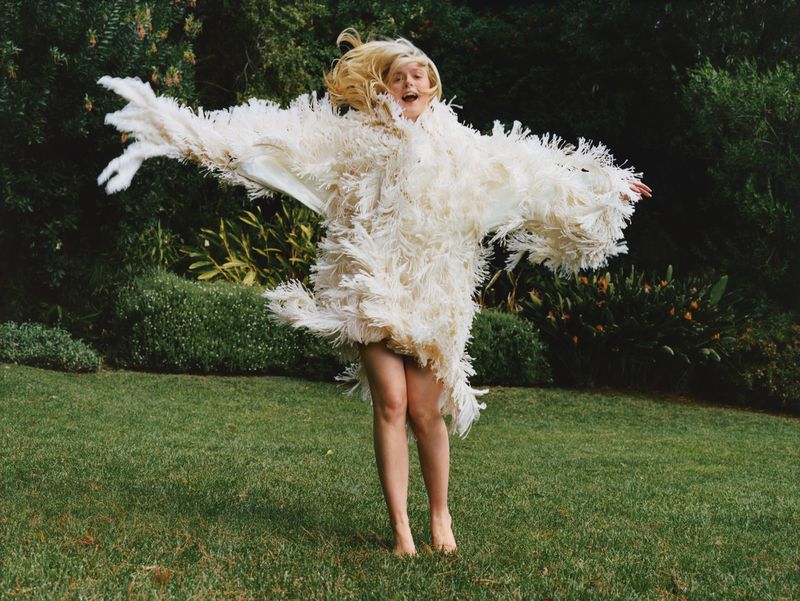 Young woman in an oversized feathered coat leaping on a green lawn in front of shrubbery, expressing joy with arms outstretched.