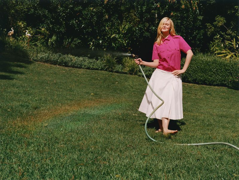 Adult female in a pink shirt and white skirt standing barefoot on a lawn holding a garden hose in a sunny outdoor setting.