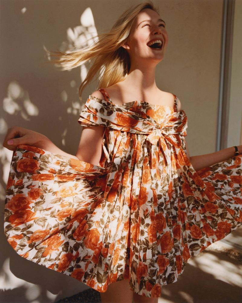 Blonde woman smiles wearing a floral print dress in natural light with shadows. The background is simple and the focus is on the subject.