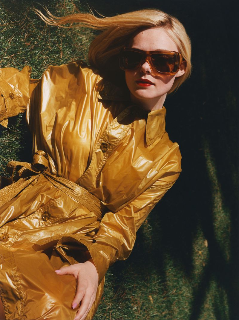Elle Fanning lies on grass wearing a shiny gold trench coat and sunglasses in an eye-level outdoor portrait, high-fashion editorial.