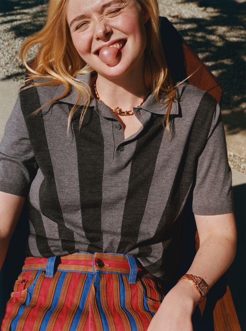 Elle Fanning poses outdoors wearing a striped polo shirt and vertically-striped pants, sticking her tongue out and smiling brightly.