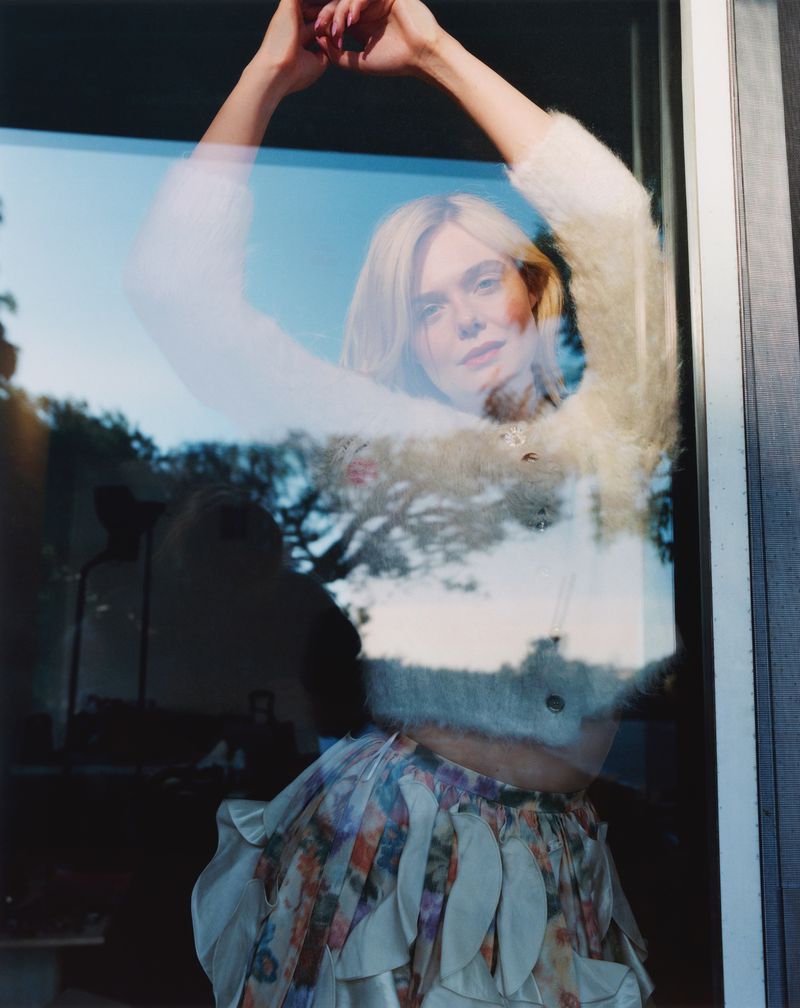 Woman with fair hair poses behind a window, wearing floral skirt and fluffy sweater, raising her arms; outdoor scene visible behind her.