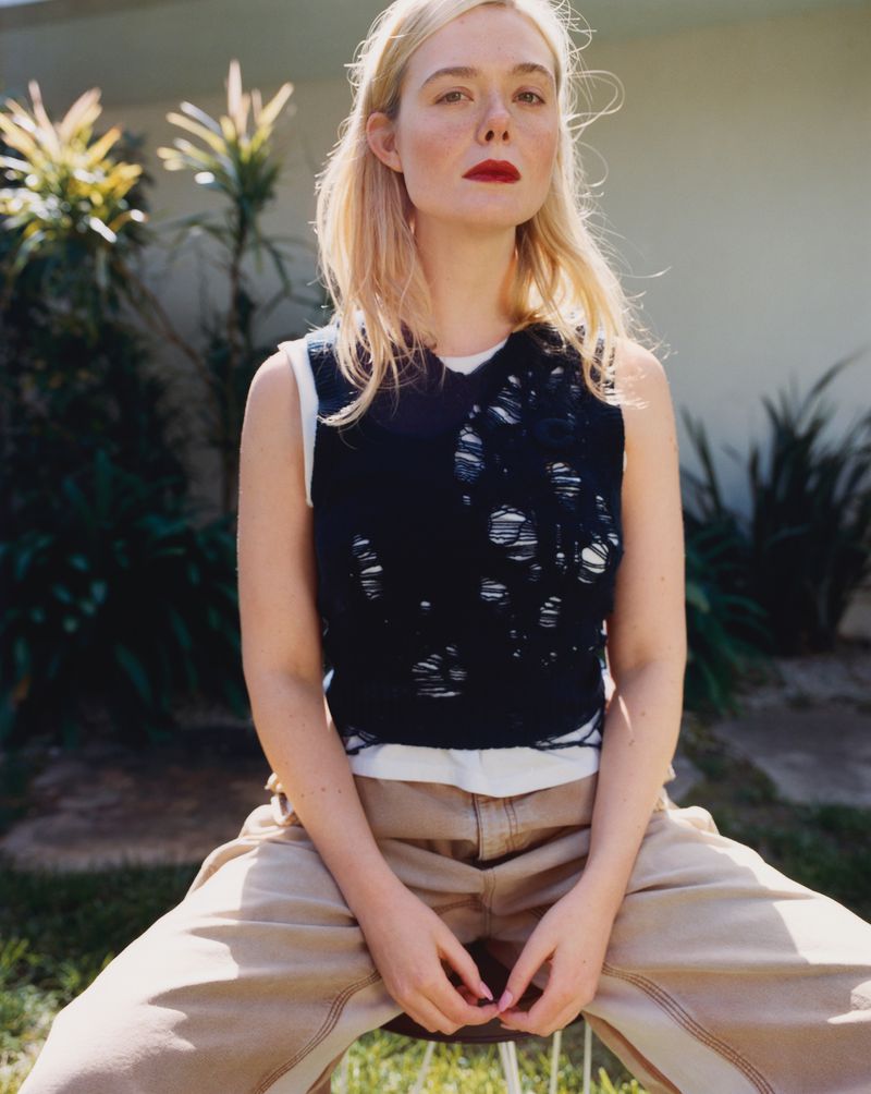 Elle Fanning poses in ripped vest top and tan pants in an outdoor setting with natural light and green vegetation in the background.