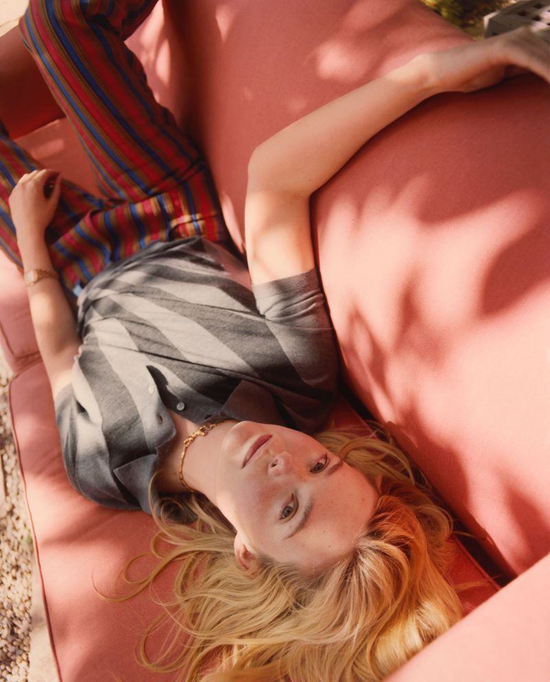 Young woman lies on pink couch outdoors wearing striped top and pants in natural light, with blonde hair, jewelry, and a wristwatch.