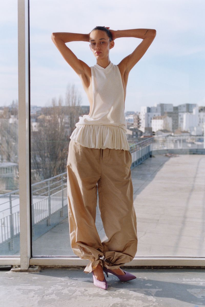 Indoor fashion shot of a slender woman in an ivory top and tan baggy pants, posing in front of a window and urban background.