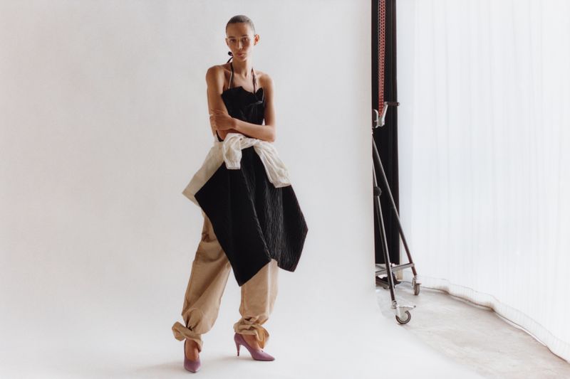 Fashion shot of a female model in a dark halter top, beige pants, and purple heels, posing in a photo studio against a white background.