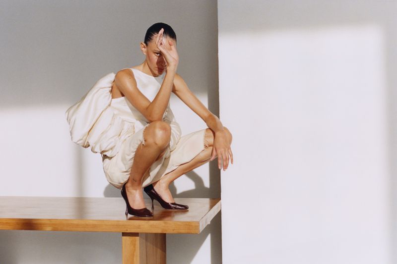A woman squats on a table, wearing a cream dress and dark heels, her hand partially covering her face in a fashion pose in studio.