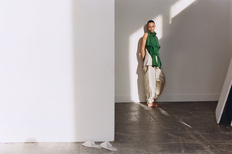 Fashionable adult female posing in a green top and cream trousers with bright window light and white walls of an indoor photo studio.