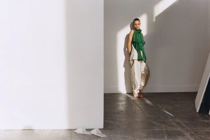 Fashionable adult female posing in a green top and cream trousers with bright window light and white walls of an indoor photo studio.