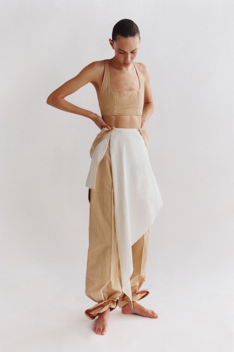 Adult female model in a tan crop top and tan and white wrap pants in a studio shot against a light background, posing with hands on hips.