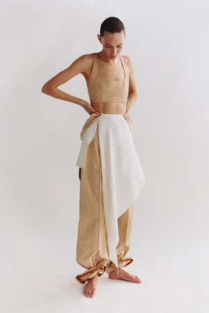 Adult female model in a tan crop top and tan and white wrap pants in a studio shot against a light background, posing with hands on hips.