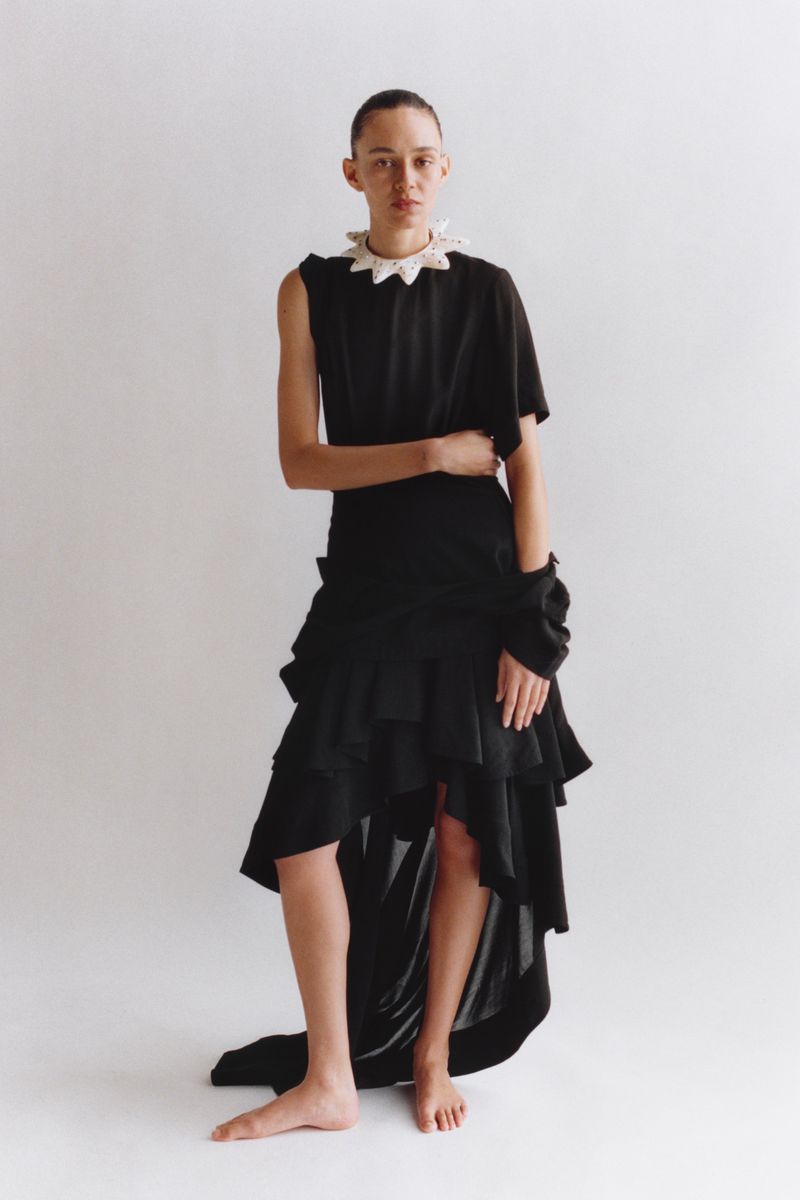 A person with short hair poses in an asymmetrical black dress with a white ruffle collar against a solid white backdrop in studio lighting.