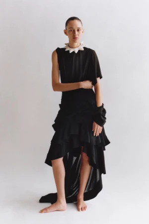 A person with short hair poses in an asymmetrical black dress with a white ruffle collar against a solid white backdrop in studio lighting.
