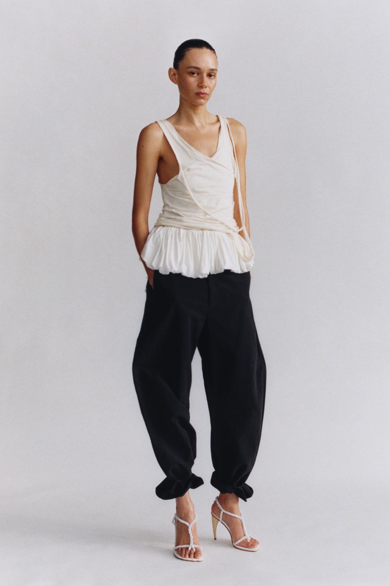 Woman in a light-colored top, black pants, and white strappy heels poses in a studio setting with a neutral background in a full shot.