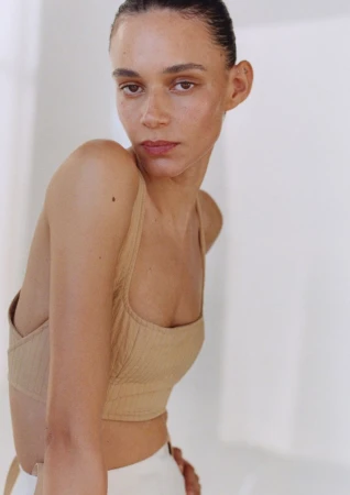 Close-up of a young adult female with brown hair pulled back, wearing a tan crop top and white pants against a white background.