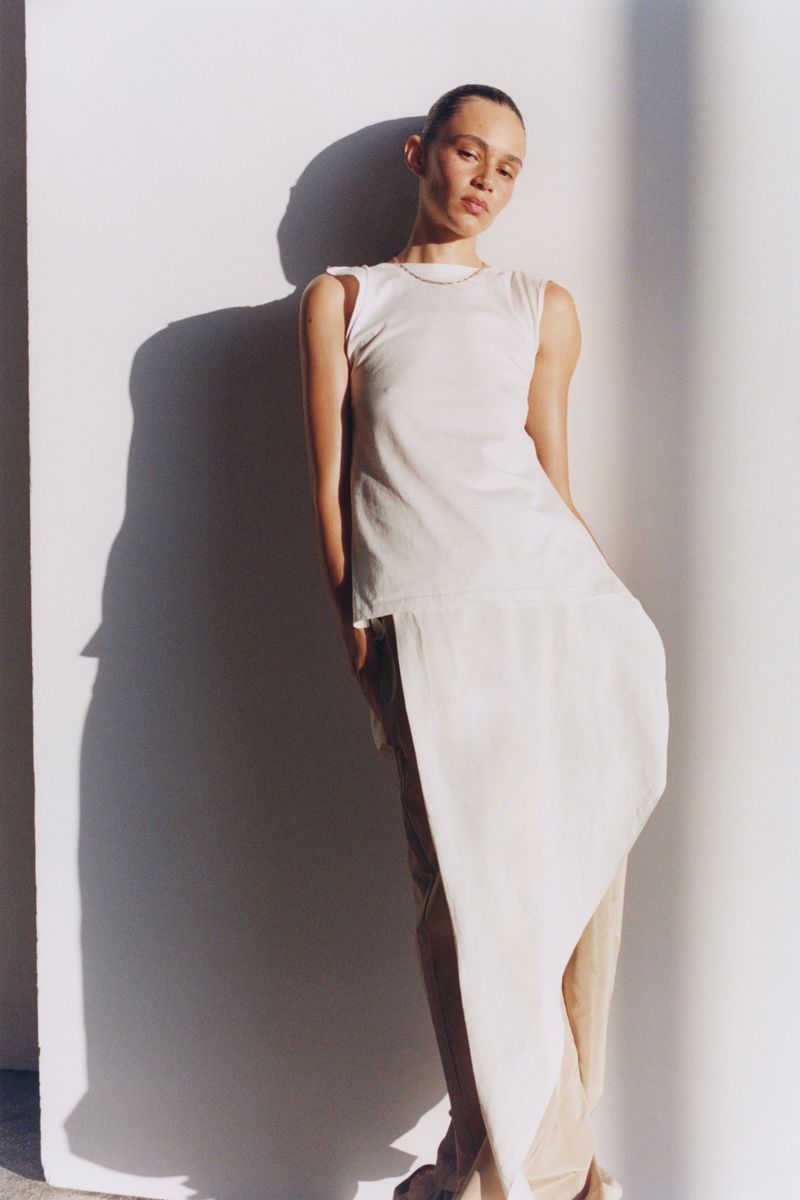 Elegant woman in an ivory dress posing against a white wall with shadow accents, captured in a fashion-style medium shot.