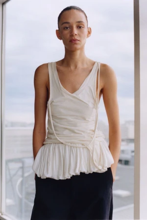 A poised woman in a light tank top and dark skirt stands indoors by a window, with soft natural light and a blurred urban background.