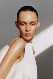 Model Binx Walton wearing a white top with sleek dark hair against a grey background with a diagonal light beam