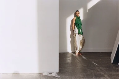 Model Binx Walton wearing a green top and trousers leaning against a white wall in Holiday Magazine editorial