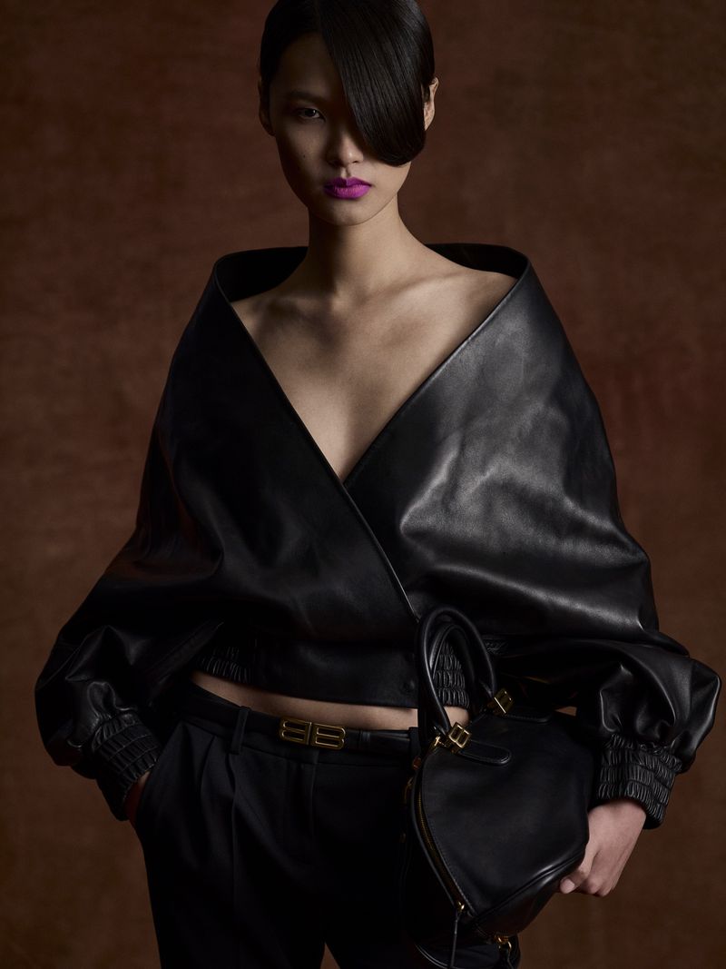 Stylish adult woman in a black leather top and pants with a purse posing against a mottled brown background in a fashion editorial shot.
