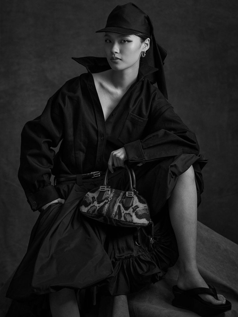 A single Asian woman is seated in a dark photo studio, wearing a black shirt, skirt, and cap, holding a leopard-print purse, looking at the camera.