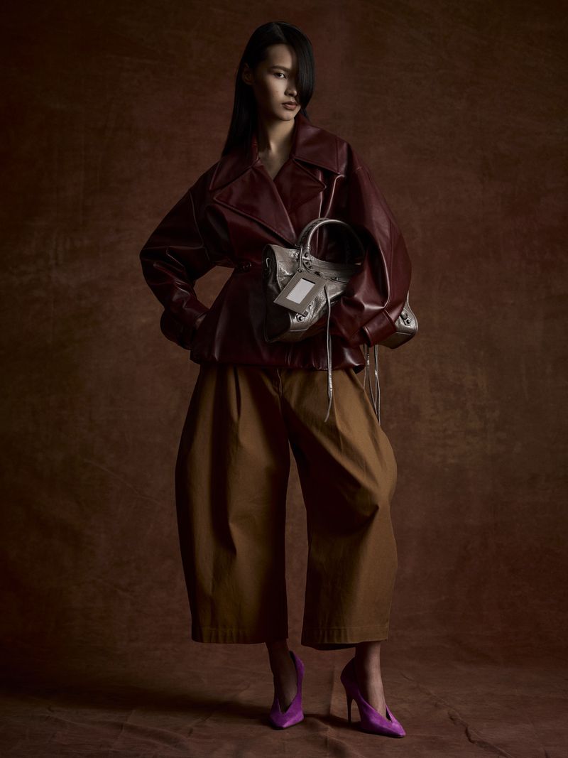 Fashionable woman posing indoors, wearing a red leather jacket, brown culottes, and purple high heels, holding a gray handbag. 