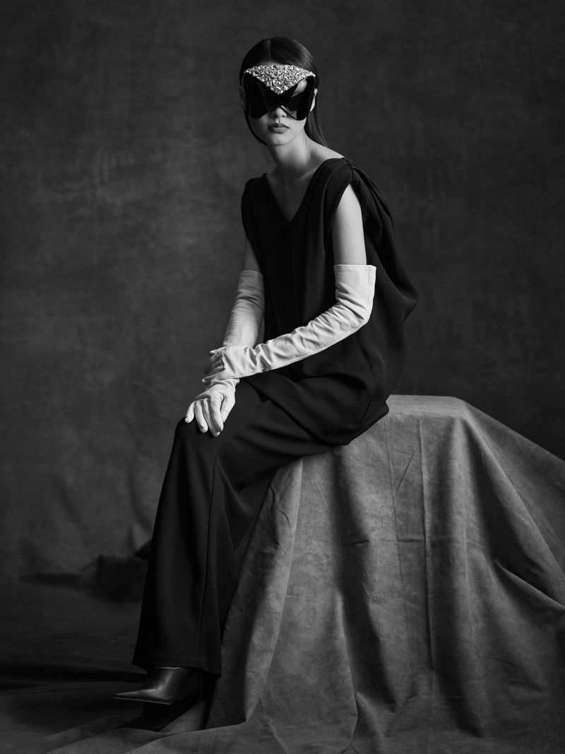 A single female model is seated wearing a black dress, gloves, mask and boots in a studio with a textured backdrop in black and white.
