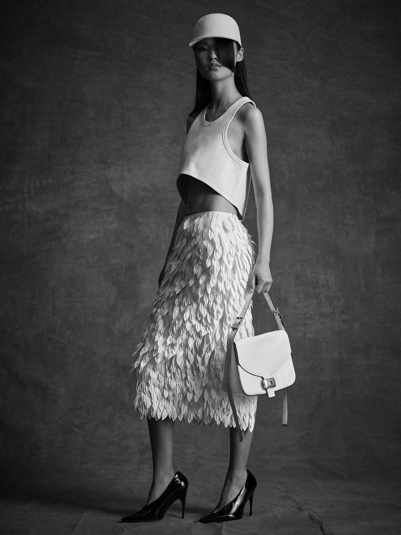 Monochrome portrait of a slim Asian woman in a cap, sleeveless top, and textured midi skirt, holding a white bag in a studio.