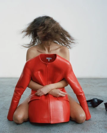 A person with flowing hair embraces a red leather Loewe jacket while kneeling against a neutral background in a fashion editorial.