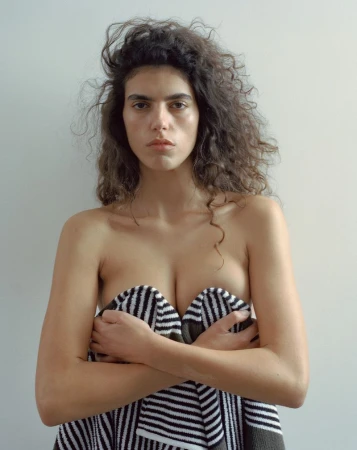 Portrait of a brunette woman with curly hair covering her chest with a striped towel against a neutral background in a studio setting.