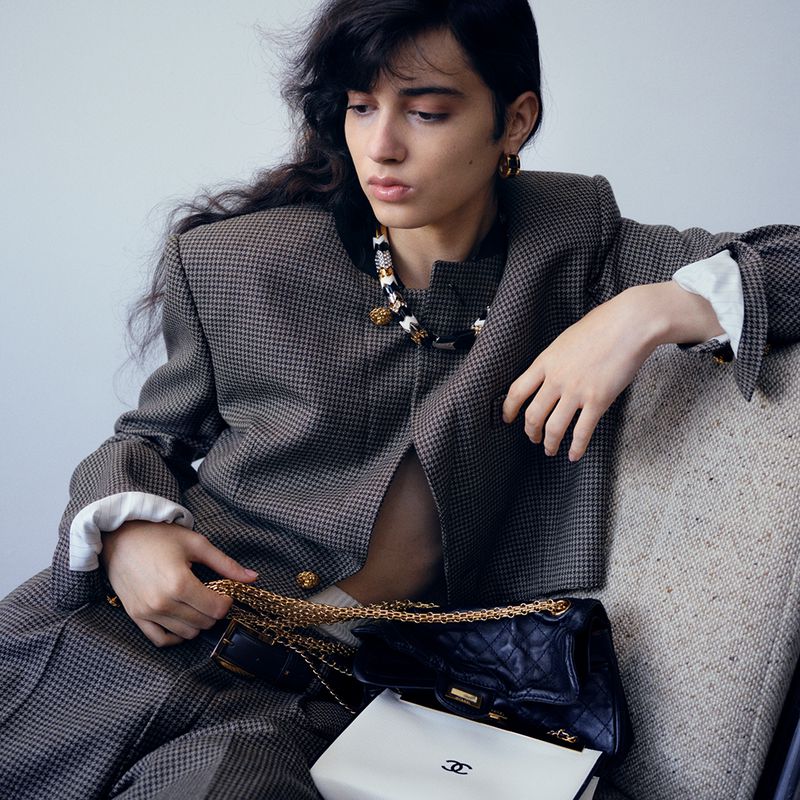 A young adult female seated in a chair, wearing a brown tweed suit with Chanel handbag and jewelry, captured in a studio setting.