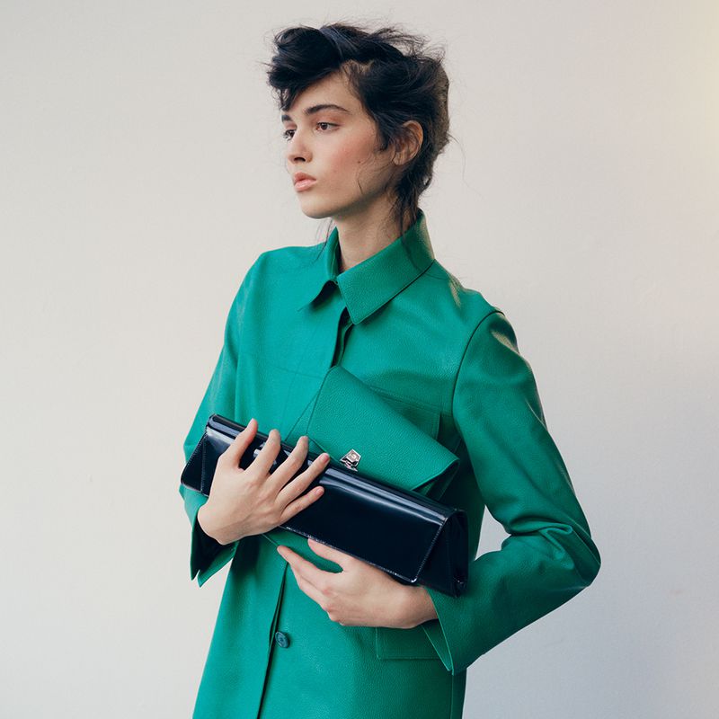 Fashionable woman in bright green coat holds two clutches, posing in front of a plain wall, showcasing style and elegance.