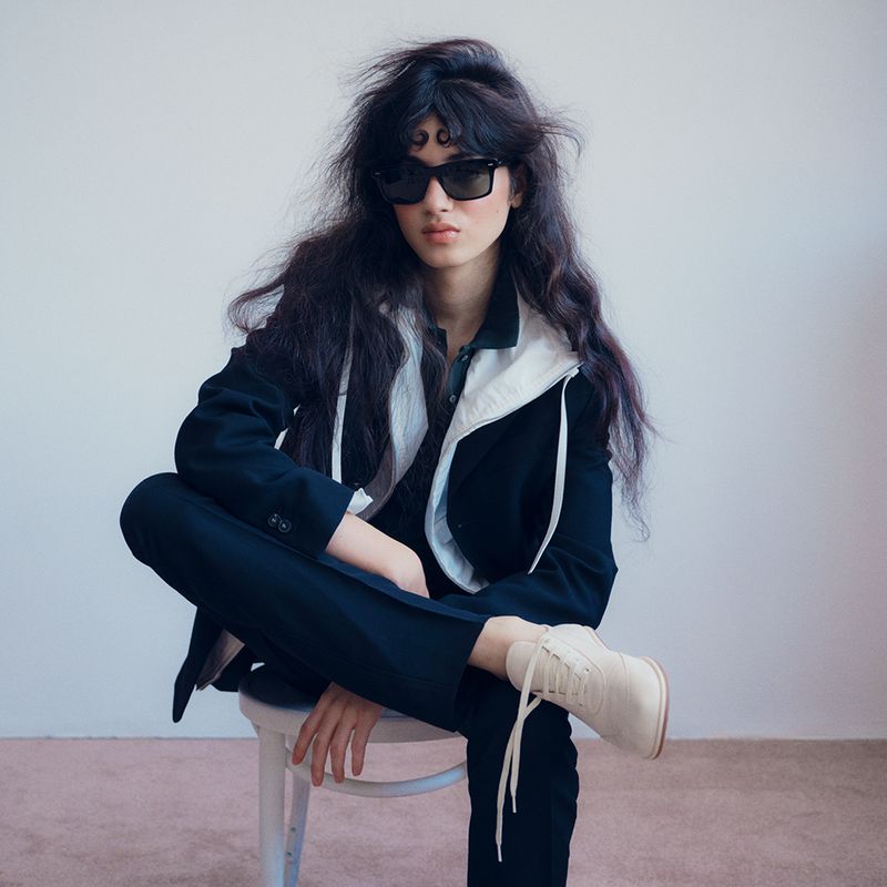 A fashionable person with dark hair and sunglasses sits on a chair in a suit and boots in a studio setting with a neutral background.