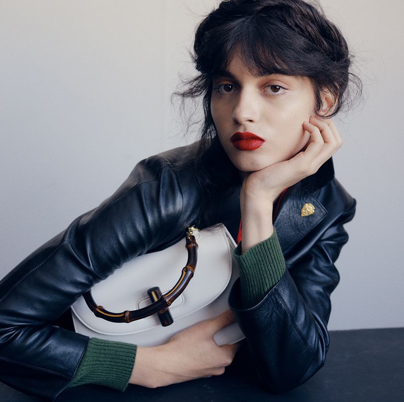 Portrait of an adult female with red lipstick and a black leather jacket, holding a white handbag with bamboo handles.