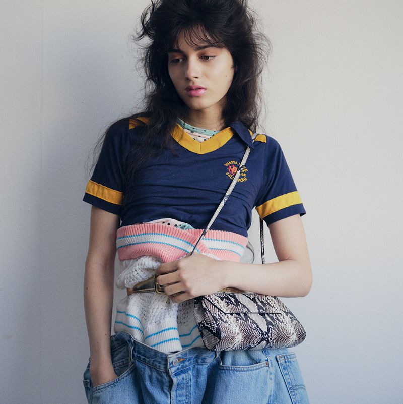 Portrait of a young woman wearing a vintage t-shirt, layered sweaters, and jeans, posing indoors in front of a neutral wall.
