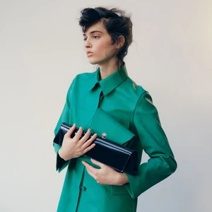 Model wearing a green coat holding a dark glossy clutch against a plain white background for The Gentlewoman.