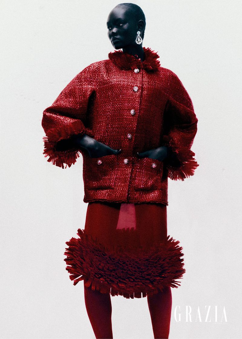 Editorial fashion shot of a black woman in a red tweed skirt suit with a ruffled collar and hemline standing on a white background.