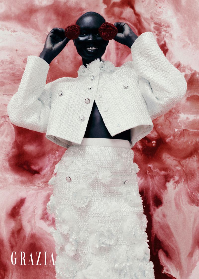 Dark-skinned woman in white floral skirt and jacket poses with red flowers near her eyes against a textured red background.