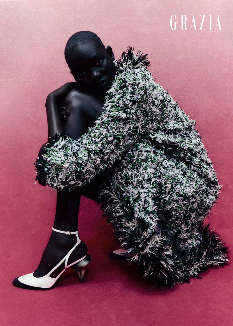 Fashion editorial shot of a black woman in a shaggy green coat, seated with one leg up, wearing white heels on a pink background.