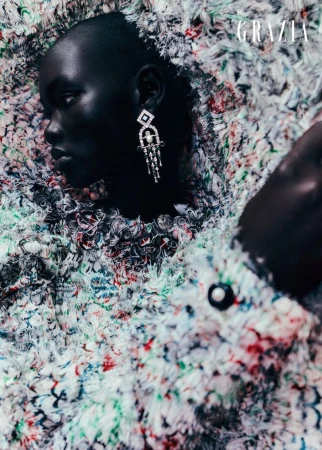 Black female model with dark complexion poses wearing an earring and a multicolor fluffy jacket in a studio for Grazia magazine.