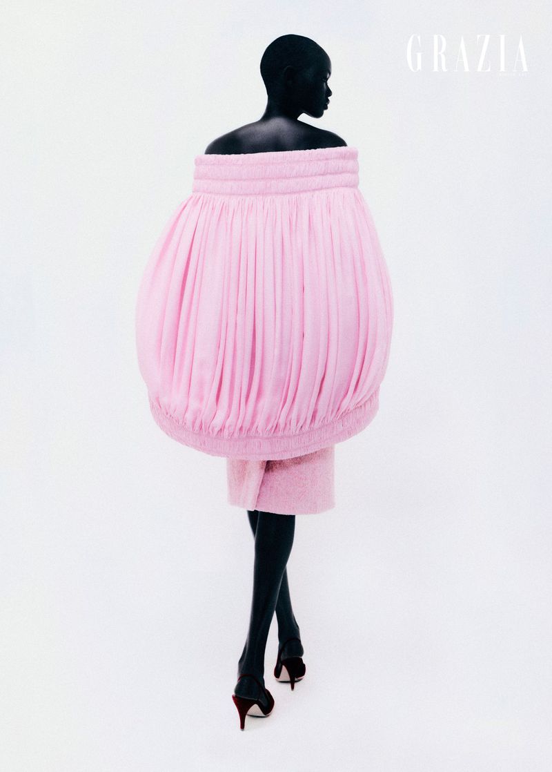 Fashion editorial shot of a black model wearing a pink sculptural dress and high heels, posing on a white backdrop with Grazia magazine logo.