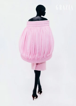Fashion editorial shot of a black model wearing a pink sculptural dress and high heels, posing on a white backdrop with Grazia magazine logo.