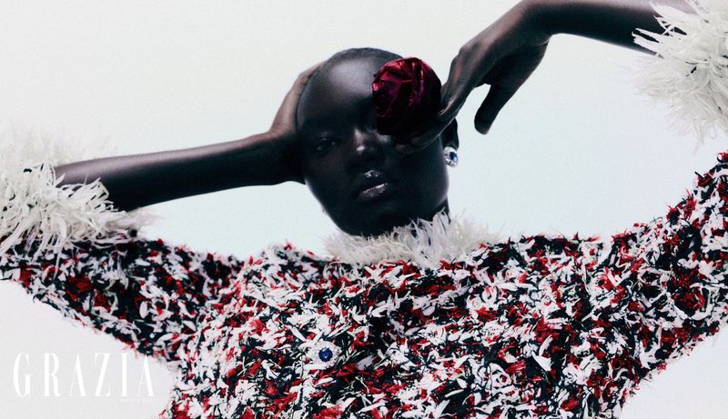 A fashion portrait of a black woman with a red flower on her head, wearing a jacket with white feather trim against a white background.