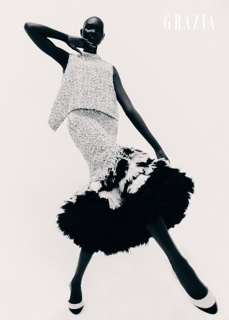 Fashion editorial shot of a Black woman in a speckled sleeveless top and skirt with feathered details, posing in front of a white background.