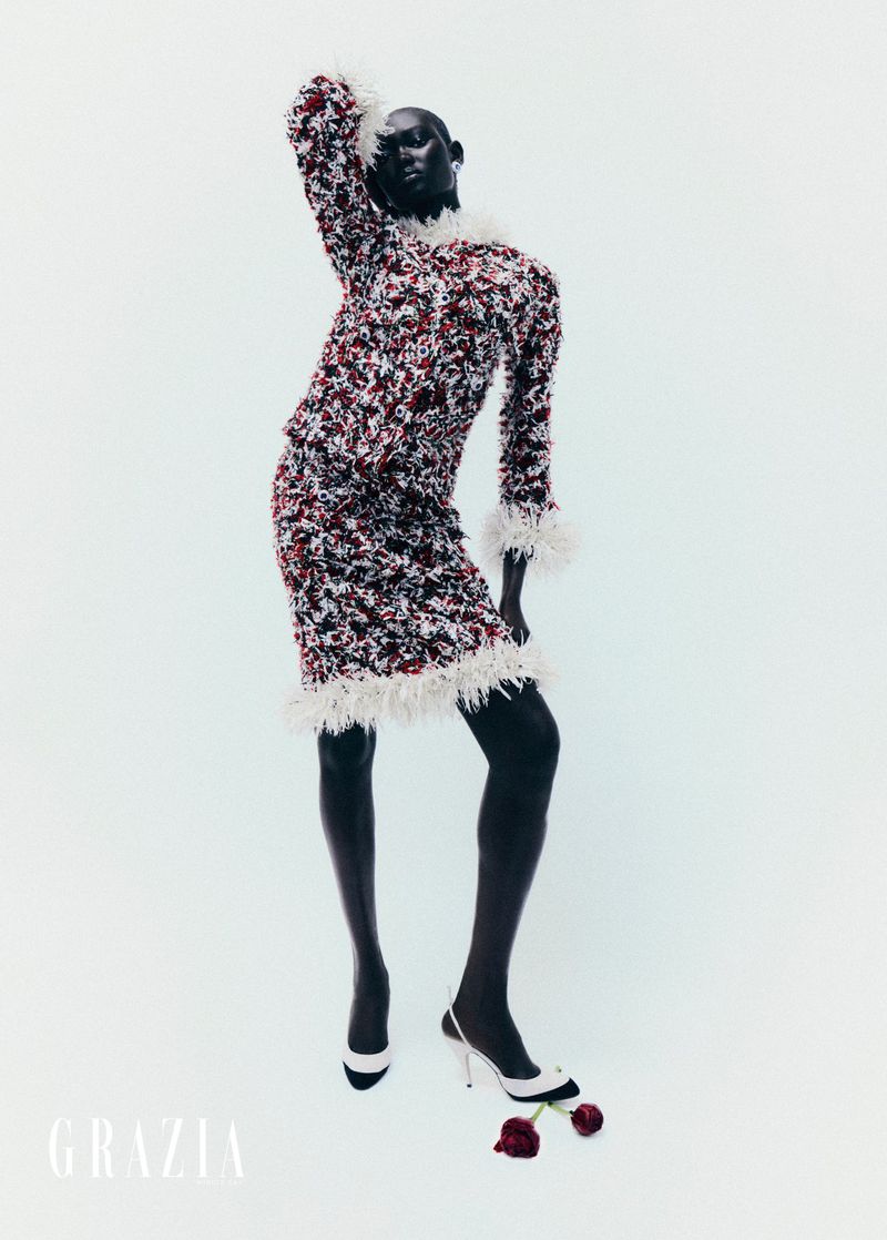 Fashion shot of dark-skinned model in patterned skirt and top, feather trim, black tights and white heels, posing against a white backdrop.