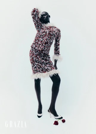 Fashion shot of dark-skinned model in patterned skirt and top, feather trim, black tights and white heels, posing against a white backdrop.